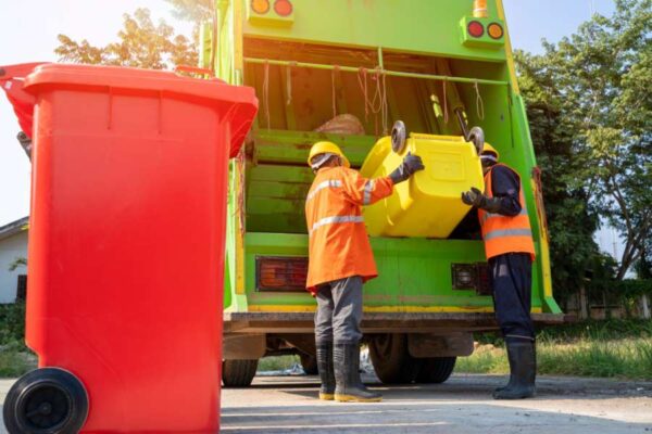 Workers from Junkisremoved loading trash bins into a green garbage truck for professional junk removal and eco-friendly waste disposal services.
