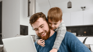 A man and a child are seated on a couch, engaging with a laptop placed on their laps